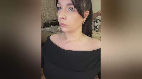 Video of a young woman with light skin, dark hair, and blue eyes, wearing a black off-the-shoulder top, in a dimly lit room with a beige textured wall and a bed in the background.