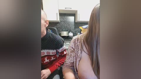 Video of a man in a red and black sweater and a woman in a patterned top, seated in a modern kitchen with white cabinets, dark countertops, and a black backsplash.