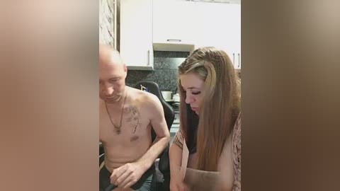 Video of a topless, tattooed bald man with a lean physique sitting on a black chair, and a young woman with long brown hair and a sleeveless top in a modern kitchen with white cabinets and a dark granite countertop.
