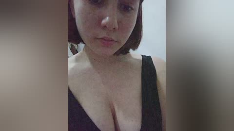 A close-up video of a fair-skinned woman with short auburn hair, wearing a black sleeveless top that accentuates her ample cleavage. The background is blurred, emphasizing her face and upper body.