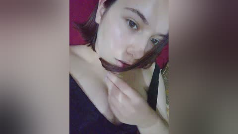 A close-up video of a young, fair-skinned woman with short, dark brown hair and a small tattoo on her arm, wearing a black off-shoulder top, with her hand touching her throat. The background is blurred, featuring a dark red surface.