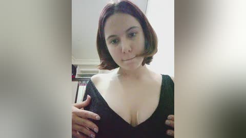 Video of a young woman with shoulder-length auburn hair, wearing a low-cut black top, standing indoors, holding her breasts, slightly smiling.