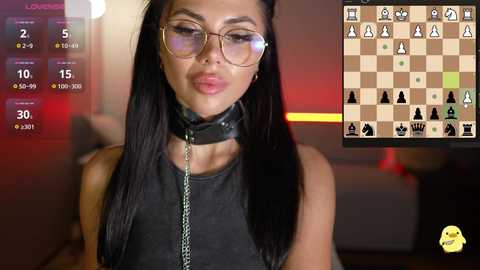 Video of a young woman with long black hair, wearing glasses, a black leather collar, and a leash, in a dimly-lit room. On the right, a chess board and digital interface.