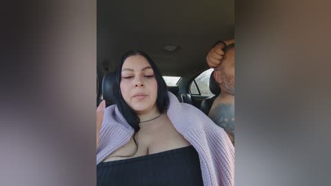 Video of a Latina woman with long black hair, wearing a black top and pink cardigan, sitting in a car, looking relaxed with a man's arm around her.