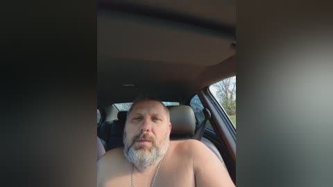 Video of a shirtless bald man with a gray beard, wearing a silver necklace, sitting in a car with tinted windows.
