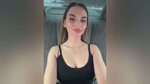 Video of a young Caucasian woman with straight brown hair, light skin, and wearing a black spaghetti-strap top. She sits in a car with gray upholstery, smiling slightly, and wearing a cross necklace.
