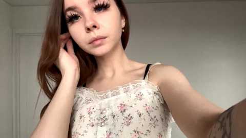 Video of a young woman with long brown hair, wearing a floral dress, looking pensive, holding her ear, in a softly lit room.