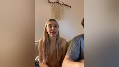 A video of a young, nude blonde woman with medium-sized breasts and a tattoo on her arm, sitting on a bed with a man in a grey shirt, in a cozy, dimly lit room with a macrame wall hanging.