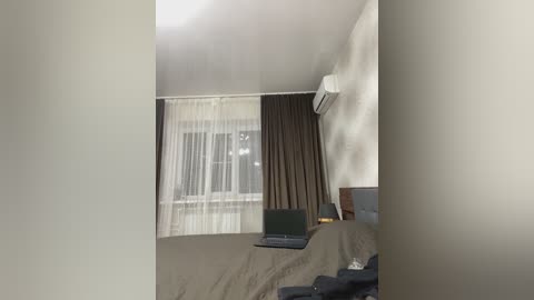 Video of a small, sparsely furnished bedroom with beige walls, a bed covered in a brown blanket, a black laptop on the bed, and a window with sheer and dark curtains.