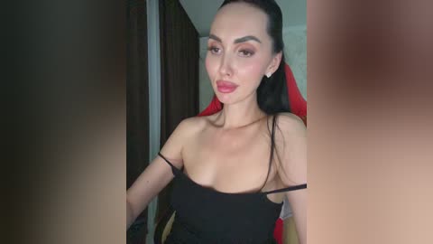 A video of a light-skinned woman with long dark hair, wearing a black strapless top, sitting indoors with a blurred background.