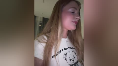 Video of a young woman with long, straight blonde hair, wearing a white T-shirt with a teddy bear graphic, standing indoors, partially obscured by a blurred, brown-toned figure.
