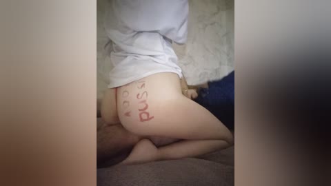 A video of a pale-skinned, nude woman with a white shirt, kneeling on a bed. She has the word \"Pussy\" written in red on her buttocks. The background shows a beige wall and a blue pillow.