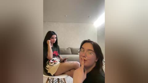 A video shows two young women in a minimalist room: one sitting on a white chair, the other standing with a finger to her lips. Both have long dark hair.