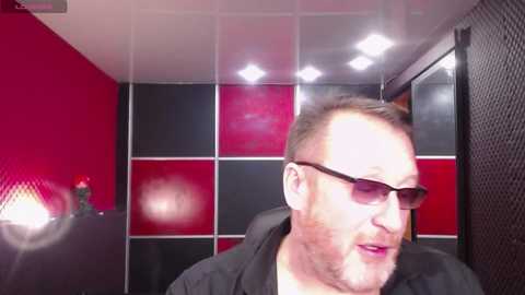 Video of a middle-aged white man with short brown hair and sunglasses, seated in a red and black checkered room with a mesh wall, wearing a black shirt.