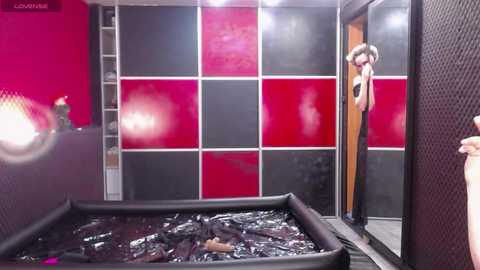 Video of a BDSM-themed room with a large black padded floor, red and black checkered walls, and a woman in a black leather outfit peeking from behind a door.