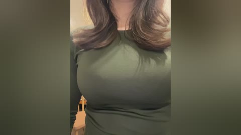 Video of a woman with medium-length, wavy brown hair, wearing a tight, dark green long-sleeve shirt that accentuates her large breasts. The background features a blurry wooden chair and a small, indistinct figure.