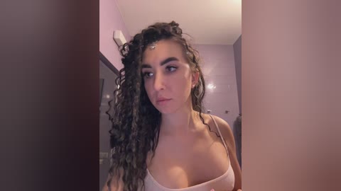 Video of a young woman with curly, dark hair, wearing a light-colored tank top, standing in a bathroom with light purple walls.