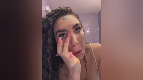 Video of a young woman with curly brown hair, wearing a white tank top, touching her forehead with a pink-painted fingernail, in a purple-lit bathroom.