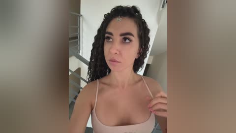 Video of a fair-skinned woman with curly dark hair, wearing a light pink spaghetti-strap top, holding her neckline. She has a delicate, small nose and a subtle makeup look. The background is a modern, minimalist interior with neutral tones and metal railings.