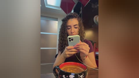 Video of a young woman with curly brown hair, holding a phone, taking a selfie in a modern room with red and white walls, and a red mug with white stars.