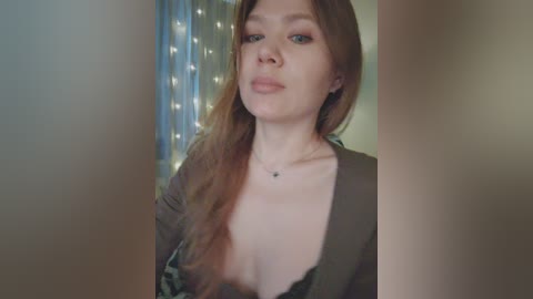 Video of a young woman with fair skin, long auburn hair, wearing a plunging neckline green top, and a delicate necklace, indoors with a blurred background featuring fairy lights.