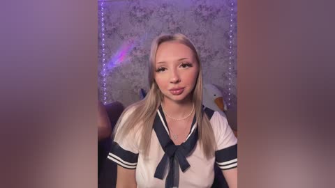 Video of a fair-skinned, blonde woman with long hair, wearing a sailor schoolgirl outfit, standing in front of a purple backdrop with fairy lights.