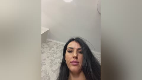 A video shows a woman with long, black hair, fair skin, and full lips, sitting on a patterned gray carpet in a minimalist room with white walls.