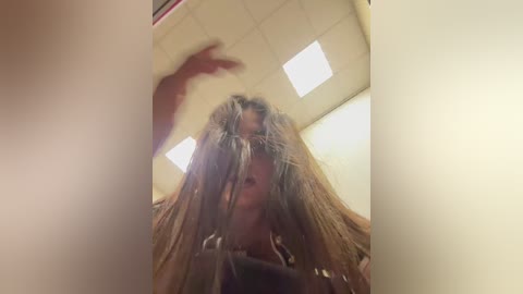 A video showing a person with messy, long brown hair, partially obscuring their face, standing in a brightly lit room with fluorescent lighting and white walls.