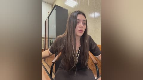 Video of a young woman with long, dark hair, wearing a black dress, seated in a wooden chair, looking surprised, in a room with beige walls and wooden accents.