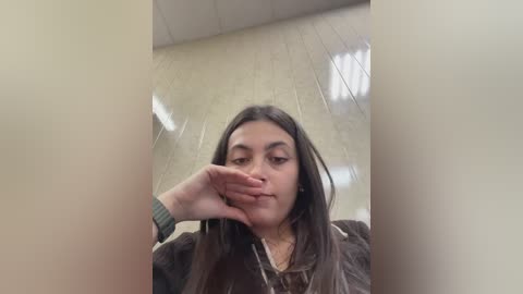 A young woman with long dark hair and fair skin, wearing a black jacket, stands in a dimly lit, beige-tiled room with fluorescent lights overhead. Her hand covers her mouth, suggesting a moment of contemplation or surprise.