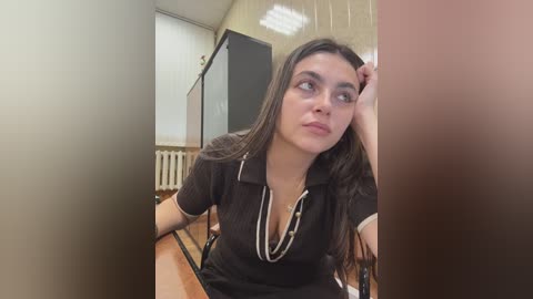 Video of a young woman with long dark hair, wearing a black polo shirt, leaning on a table in a classroom with beige walls, a radiator, and a blackboard.