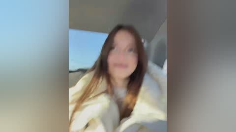 A blurry video of a young woman with long brown hair and fair skin, wearing a white robe, seated indoors with a bed and window in the background.