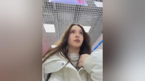 Video of a young woman with long brown hair, wearing a white coat and a gray sweater, looking up in a subway car with tiled ceiling and fluorescent lights.