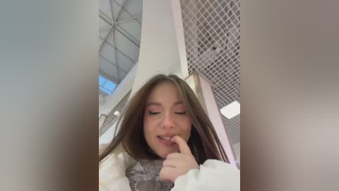 Video of a young woman with long brown hair, wearing a white sweater and gray scarf, making a peace sign with her finger. Background features a modern building with metallic accents and geometric patterns.