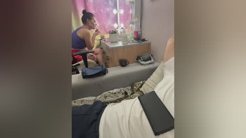 A video of a woman in a purple tank top and red shorts sitting on a bench, watching a man lying in bed with a tablet in hand.