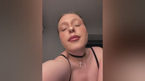 Video of a fair-skinned, light-haired woman with closed eyes, wearing a black choker and a pink dress, in a blurred room.