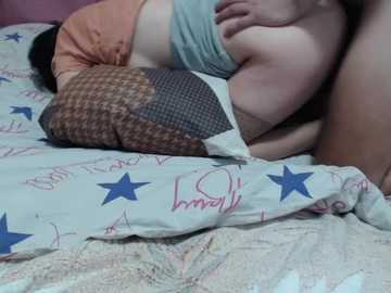 Video of a large, plus-sized woman lying on a bed with a star-patterned duvet, partially covered by a plaid blanket. She is wearing a grey bra and a blue shirt, with her right hand on her breast.