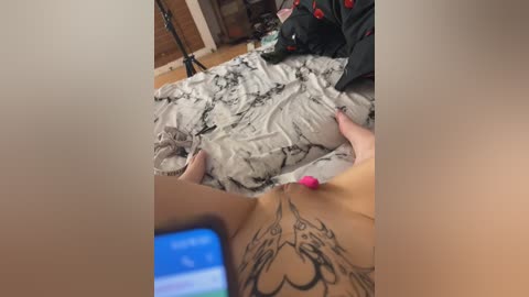 A video shows a person with light skin and tattoos lying on a bed with black and white marble-patterned sheets. A smartphone with a blue screen is visible in the foreground.