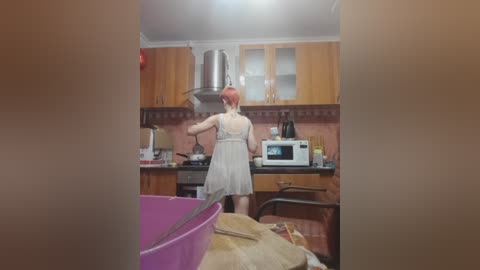 Video of a woman with red hair in a white dress cooking in a small, cluttered kitchen with wooden cabinets, a microwave, and a stove.