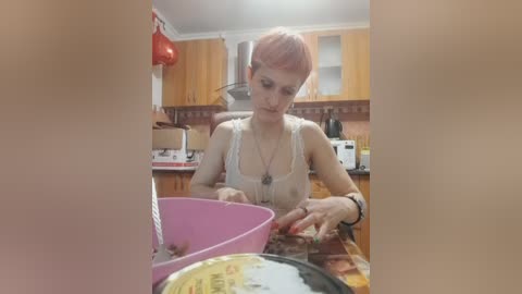 Video of a fair-skinned woman with short pink hair, wearing a beige tank top, mixing cake batter in a pink bowl in a kitchen with wooden cabinets and red hanging pot holders.