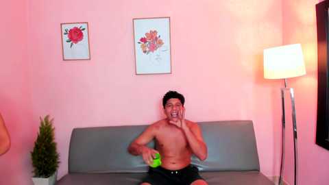 Video of a shirtless, fit young man with dark hair, holding a green apple, sitting on a gray couch against a pink wall. Artwork and a floor lamp are visible in the background.