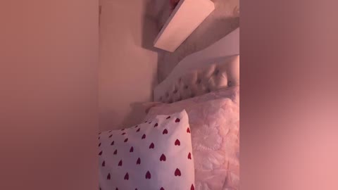 Video of a small, dimly lit bedroom with a white bed covered in a quilt featuring red heart patterns. The room has a plain, beige wall with a white shelf above the bed.