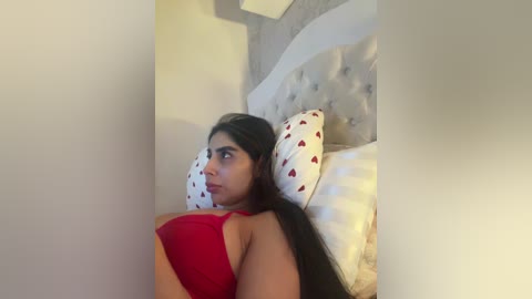 Video of a young woman with medium skin tone, dark hair, and a red bra, lying on a white bed with a heart-patterned pillow. The background is a white wall with a modern, tufted headboard.