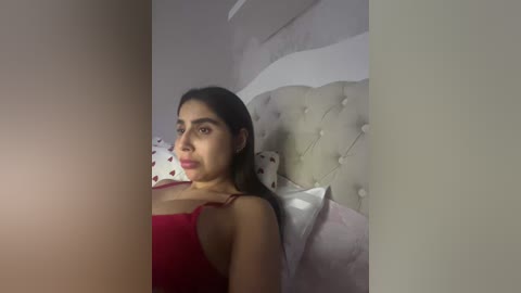Video of a young woman with medium brown skin, dark hair, and a slender build, wearing a red tank top, lying on a bed with a tufted headboard.