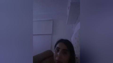 A dimly lit video of a woman with long dark hair, sitting in a small, dimly lit room. She has a contemplative expression, looking downwards. The background features a white wall and a partially visible white door.