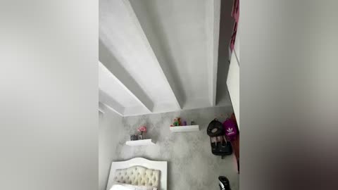 A video of a minimalist bedroom with a high ceiling, white walls, and a gray concrete floor. A bed with a white headboard and a beige quilt is visible, along with a white nightstand holding a pink flower vase. A black bag and a red object are placed against the wall.