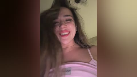 Video of a smiling Asian woman with long dark hair, wearing a pink tank top, in a dimly lit room with beige walls.