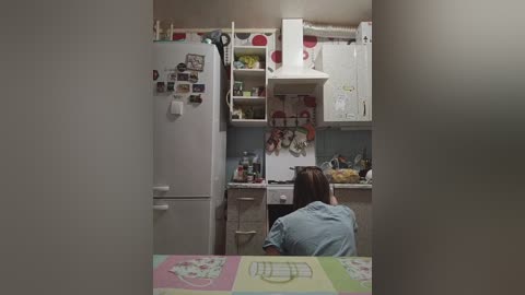 A video of a small, cluttered kitchen with white cabinets, a white fridge, and a woman with brown hair sitting at a table.