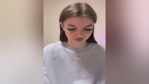 Video of a young woman with long brown hair, fair skin, wearing a light gray hoodie, standing in a minimalist room with beige and pink walls.