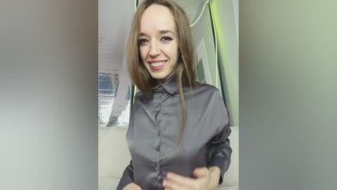 Video of a smiling Caucasian woman with shoulder-length light brown hair, wearing a shiny gray blouse. She stands indoors, near a window with a green light and a blurred, white wall in the background.